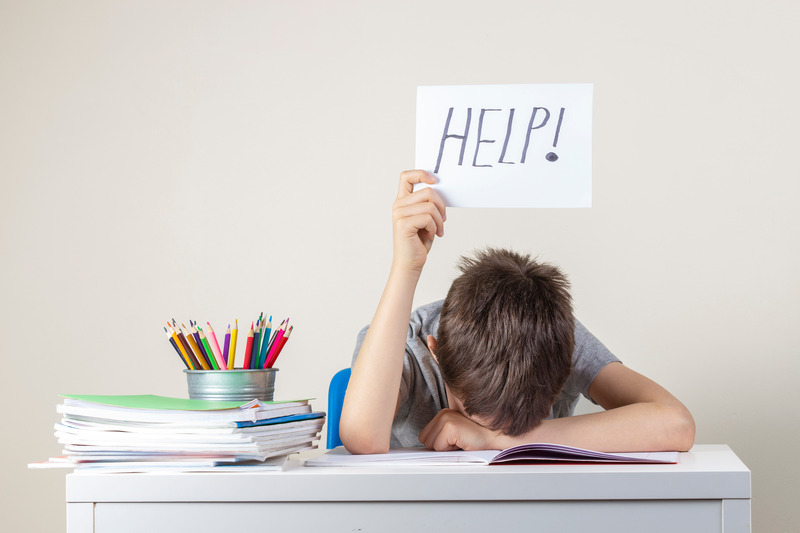 A child sits at a cluttered desk, head down, holding a 'HELP!' sign.
